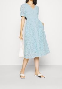 Světle modré šaty s krátkými nafouklými rukávy a výstřihem do V. Obsahují květinový vyšívaný vzor, řasený pas a splývavou sukni. V doprovodu bílých doplňků.