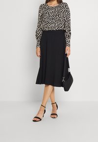 Schwarzer Rock kombiniert mit einem langärmeligen Oberteil mit einem beige- und cremefarbenen Punktmuster. Die Accessoires umfassen eine schwarze Handtasche und schwarze, schlichte High Heels.