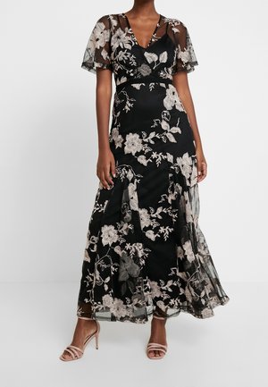 Zwarte maxi-jurk met bloemenprint en doorschijnende, zwierige mouwen, voorzien van verfijnde beige borduursels en een aansluitende taille. Een split zorgt voor meer beweging.