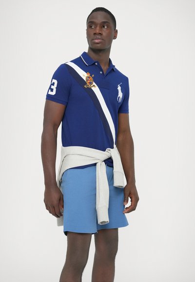 Tengerészkék galléros póló fehér átlós csíkkal, hímzett logóval és a 3-as számmal; világoskék rövidnadrág; szürke pulóver a deréknál megkötve.