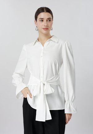 Femeie care poartă o bluză albă cu mâneci lungi, cu nasturi și cordon în talie, și pantaloni negri, stând cu o mână în buzunar, privind spre dreapta.