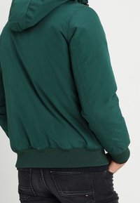 Giacca imbottita verde con cappuccio, polsini e orlo a coste. Texture liscia con semplici dettagli di cucitura, progettata per calore e comfort.