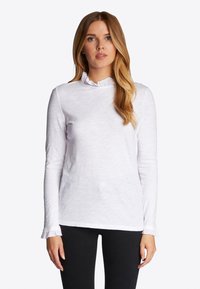 Top blanco de manga larga con cuello y puños con volantes, tejido ligero y texturizado, y un corte relajado, combinado con pantalones negros.