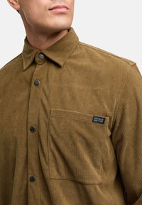 Camisa de pana marrón con un collar estructurado, cierre frontal de botones y un bolsillo en el pecho que presenta una etiqueta rectangular. Textura de canalé sutil.