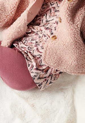 Zbliżenie na warstwowe ubrania pokazujące różowy pluszowy kurtkę, wzorzystą bluzę z sznurkiem oraz liliowe legginsy na białej puszystej powierzchni.