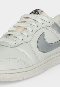 Λευκό αθλητικό παπούτσι Nike Air με ασημένιο σήμα, λευκά κορδόνια και υφασμένο καουτσούκ στην σόλα, εμφανιζόμενο από κοντινή πλευρική γωνία.