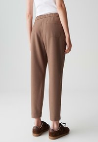 Personne portant un pantalon brun cintré avec une poche arrière et des baskets brunes, se tenant devant un fond gris clair uni.