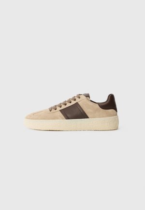 Baskets en suédine beige avec des accents en cuir brun foncé, lacets plats et semelle crème texturée. Détails de rayures sur le côté ajoutent un élément de design.