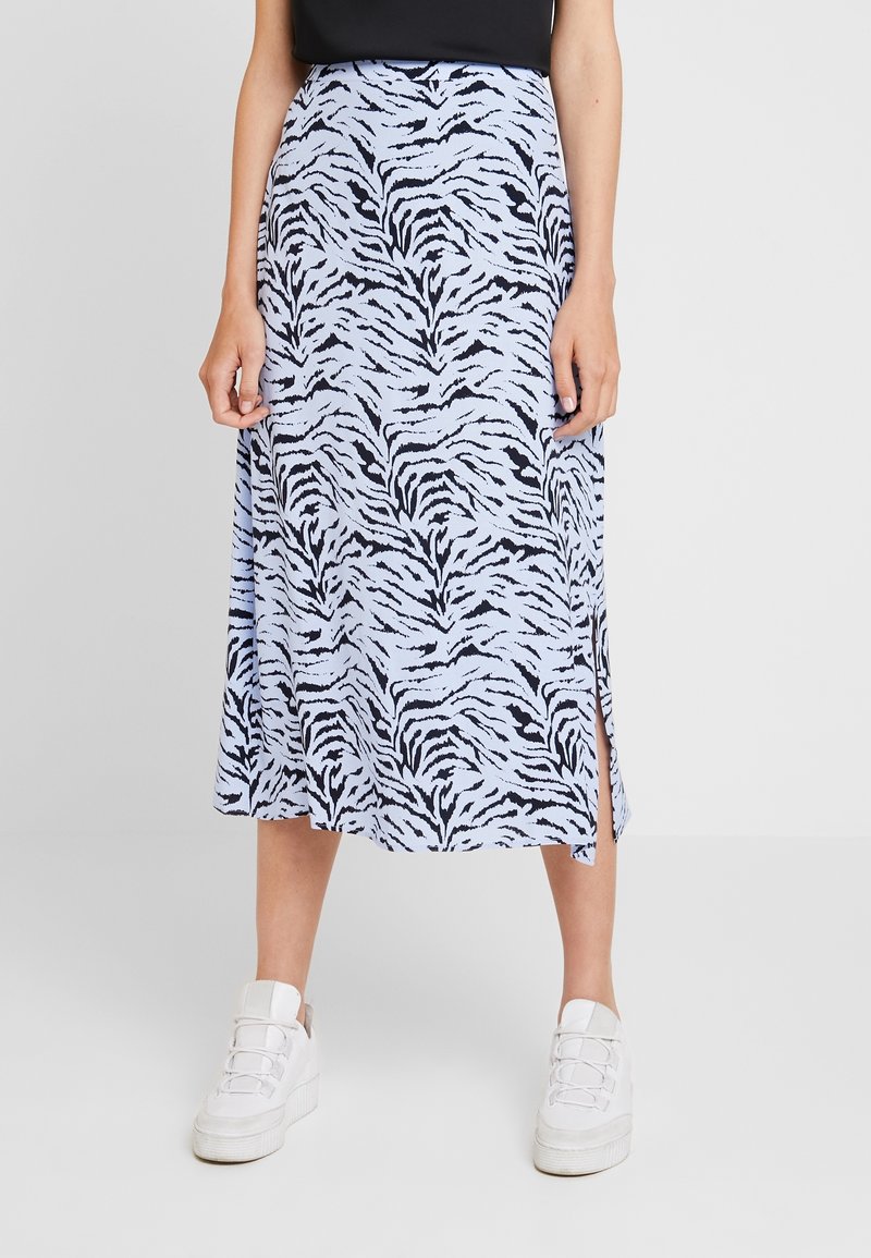 Femme portant une jupe midi bleu clair avec imprimé zèbre noir, fente latérale, baskets blanches et un haut noir, photographiée de la taille jusqu'aux pieds.