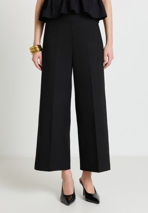 Donna che indossa pantaloni neri a gamba larga e corti, tacchi alti neri e numerosi braccialetti dorati al polso sinistro, su uno sfondo semplice.