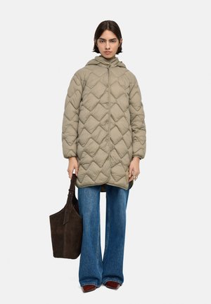 Kobieta w beżowym pikowanym płaszczu z kapturem, niebieskich szerokich spodniach dżinsowych, brązowych butach, trzymająca dużą ciemnobrązową torbę typu shopper, stoi na białym tle.