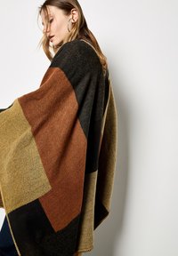 Wollen poncho met een geometrisch patroon in bruin, zwart en mosterdkleur. Afgewerkt met een decoratieve gestikte rand. Zachte textuur en warm ontwerp.