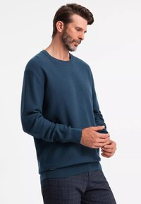 Maglione blu scuro da uomo, con motivo a maglia testurizzata, scollatura a girocollo, polsini e orlo a coste, abbinato a pantaloni fantasia, mani posizionate in vita.