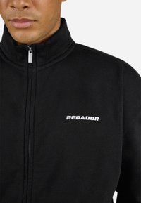 Zwarte zip-up jas van zachte stof, met een hoge kraag en het witte "PEGADOR" logo op de linkerborst. Glad textuur.