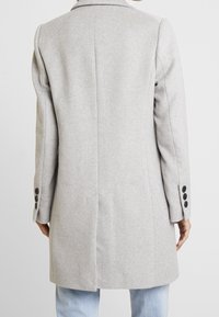 Grey wool coat with tailored fit, notched collar, and pronounced shoulder seams. Features black button accents on cuffs. Smooth texture.