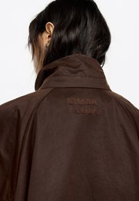Chaqueta marrón con cuello alto y el logo bordado "Bimba y Lola". Textura suave con un brillo sutil y detalles de costura limpios.