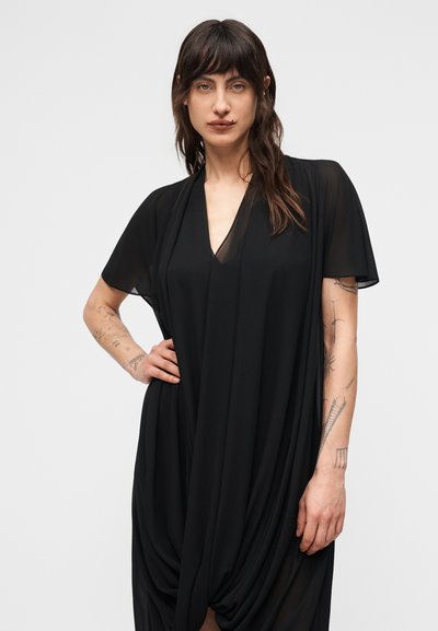 Άτομο με μακριά σκούρα μαλλιά και τατουάζ, που φοράει ένα φαρδύ μαύρο φόρεμα με κοντά διαφανή μανίκια, στέκεται μπροστά σε απλό φόντο.