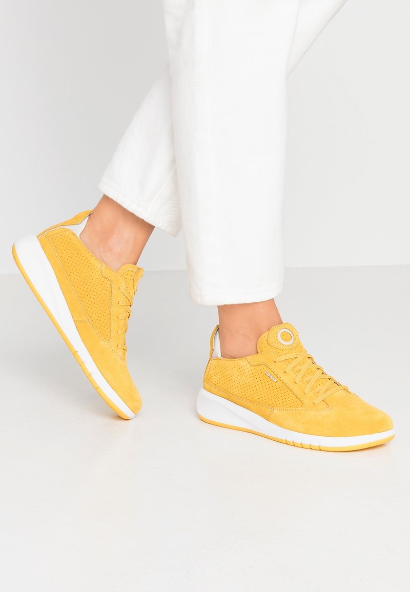 Personne portant des baskets en suède jaune avec des semelles blanches et des détails perforés, associées à un pantalon blanc, sur un fond uni.