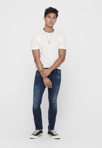Weißes, strukturiertes T-Shirt, eng anliegende blaue Jeans, schwarze Sneakers mit weißen Sohlen, stehende Pose, minimale Accessoires. Schlichter Hintergrund.