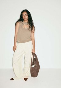 Femme aux longs cheveux bouclés portant un haut beige sans manches et un pantalon large crème, tenant un grand sac à main marron en daim.