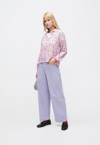 Camicia rosa con motivo floreale, bottoni sul davanti e maniche con polsini, abbinata a pantaloni larghi in velluto a coste color lavanda e scarpe nere.