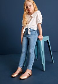 Weißes Kurzarm-Bluse mit gerüschtem Kragen und Band vorne, enganliegende Jeans aus Denim und metallic bronzefarbene Mary-Jane-Schuhe. Blauer Hintergrund.