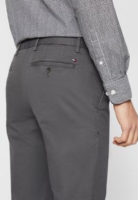 Šedé bavlněné kalhoty se slim fit střihem, s zadní kapsou na knoflík a malou značkovou štítkou. Materiál je hladký.