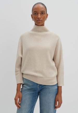 Beige Rollkragenpullover mit lockerer Passform und langen Ärmeln, aus einem weichen, strukturierten Material. Kombiniert mit hellblauen Jeans.