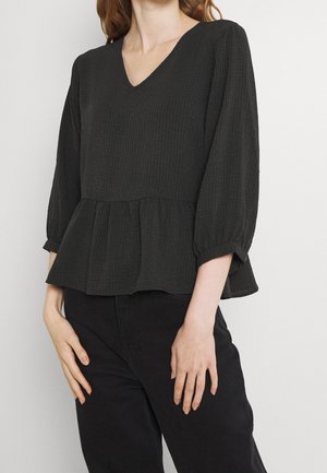 Kobieta ubrana w czarną teksturowaną bluzkę z dekoltem w serek, rękawami 3/4 i peplum, połączoną z czarnymi spodniami na jednolitym tle.
