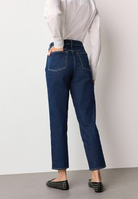 Tamsiai mėlyni džinsai su aukštu juosmeniu, tiesiomis kojomis ir penkių kišenių dizainu. Dėvimi su balta marškinių tipo palaidine ir juodais butais su smaigaliais.