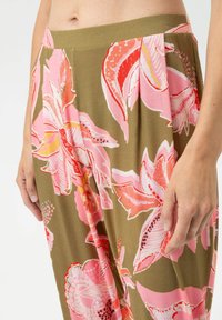 Close-up van een persoon die olijfgroene broek met grote roze en rode bloemenprints draagt, waarbij een deel van de taille, heupen en handen zichtbaar is.