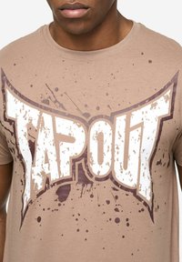Bež majica s istaknutim bijelim i smeđim logotipom "TAPOUT" s uzorcima prskanja; mekani materijal, dizajn s okruglim izrezom.