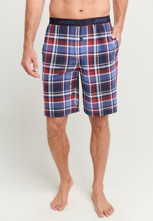 Mann trägt rot-navy-weiße karierte Shorts bis zur Mitte des Oberschenkels mit schwarzem elastischem Bund mit der Aufschrift „Jockey“ und steht barfuß vor hellem Hintergrund.