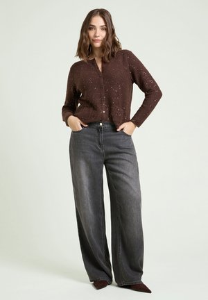Woman with shoulder-length brown hair stands with hands in pockets wearing a brown speckled cardigan and loose gray jeans.
