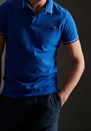 Niebieska koszulka polo wykonana z teksturowanej tkaniny, z kołnierzykiem w kontrastowe paski i logo na piersi. Nosi się ją na co dzień w połączeniu z ciemnymi spodniami.
