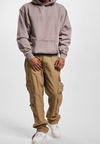 Persona vestida con un sudadera con capucha malva, pantalones cargo color beige con múltiples bolsillos y zapatillas blancas, de pie contra un fondo liso.