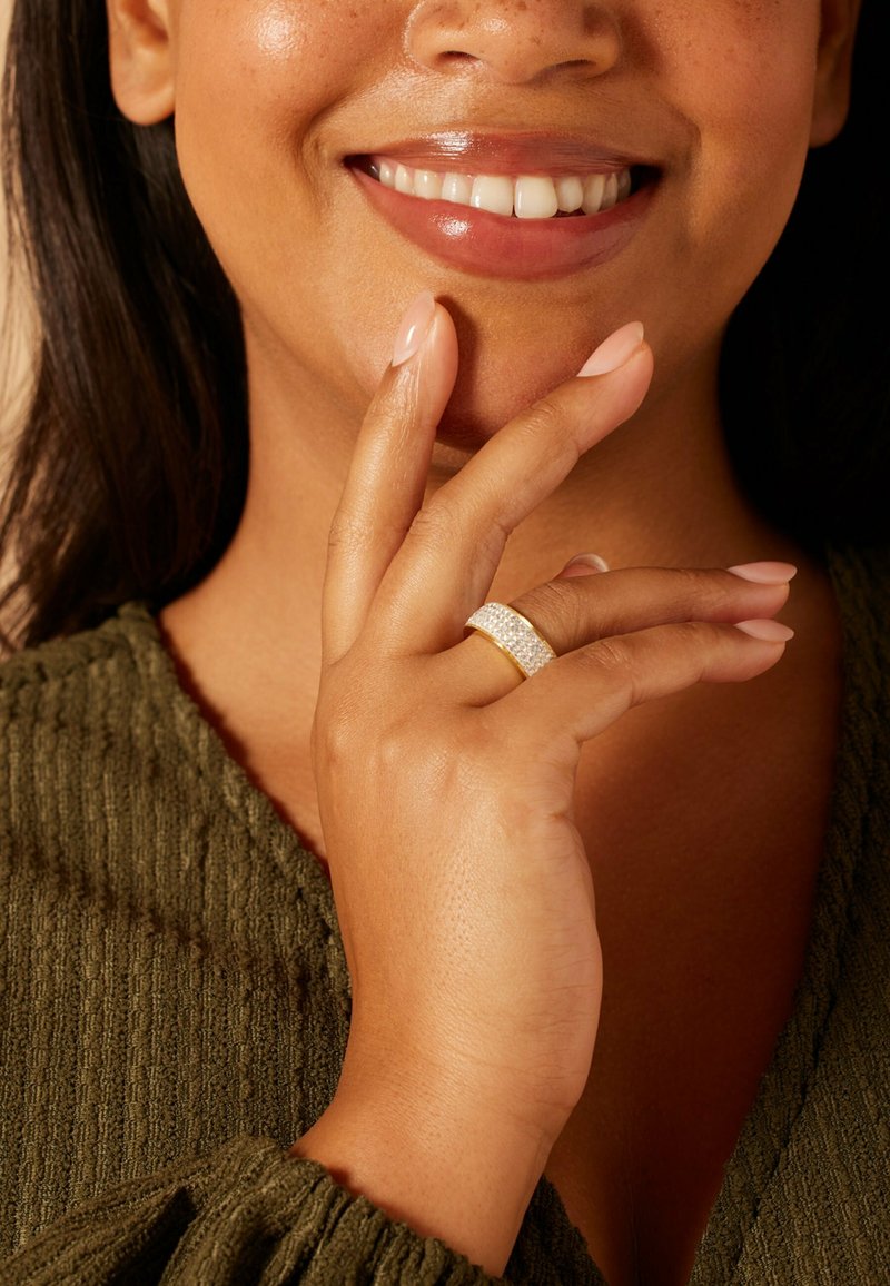 Gold ring with white gemstone accents worn on a hand with light nail polish, set against a textured olive green fabric background.