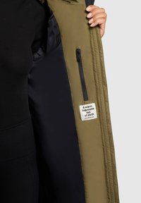 Olivgrön ytterjacka med svart foder, med en ficka med dragkedja och en tygetikett som säger "En varm kram gjord av tyg."