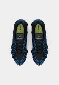Par de ténis Nike Shox em preto e azul com palmilhas verdes, vistos de cima sobre um fundo branco.