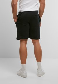 Shorts noirs avec une taille élastique, des poches latérales et une poche arrière, fabriqués à partir d'un tissu lisse. Portés avec des baskets blanches.