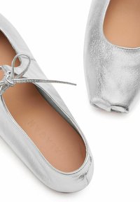 Sølv metallic ballet flats med afrundet tå og sløjfedetalje på venstre sko, tan indvendig foring, vist på hvid baggrund.