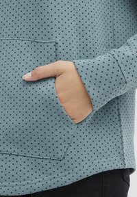 Svetlo modra sweatshirt s majhnimi črnimi pikami, ki ima sprednji žep in manšete na rokavih. Mehak material in sproščen kroj.