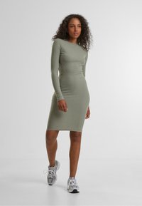 Abito a maniche lunghe, a coste, di un verde oliva tenue, con una silhouette aderente e lunghezza al ginocchio, abbinato a scarpe da ginnastica grigie.
