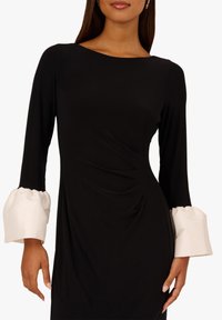 Czarna sukienka z długimi rękawami, z białymi falbaniastymi mankietami i marszczonym fasonem w talii, uszyta z gładkiego, elastycznego materiału.