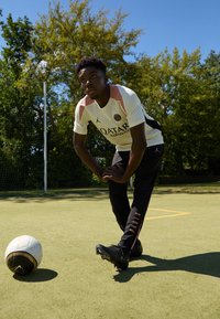 Młody mężczyzna w strojach sportowych rozciąga się na boisku piłkarskim na świeżym powietrzu, obok piłki, pod czystym, niebieskim niebem, z drzewami w tle.