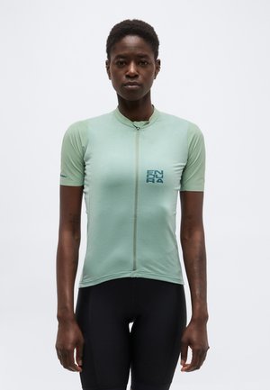 Kvinde iført en lysegrøn cykeltrøje med korte ærmer og lynlås foran samt sorte cykelshorts, stående mod en ensfarvet baggrund.