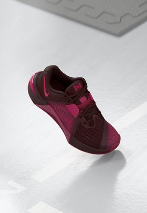 Zapatilla deportiva de color marrón oscuro con acentos en rosa. Presenta una parte superior de malla, punta redondeada y suela texturizada. Cordones y logo exhibidos de manera prominente.