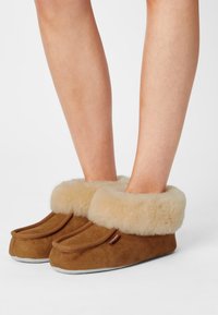 Chaussons en suède marron avec un large revers en fourrure beige, dotés d'un bout rond et d'une semelle grise contrastante. Texture douce et design simple.