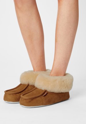 Bruna mocka tofflor med en bred klädd kant i tan-färg, med rund tå och kontrasterande grå sula. Mjuk textur och enkel design.
