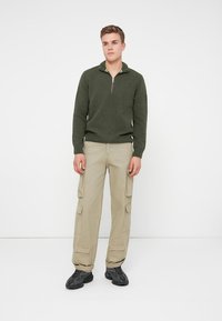 Tmavozelený pletený sveter na zips, béžové cargo nohavice s bočnými vreckami a čierne tenisky. Model stojí na bielom pozadí.